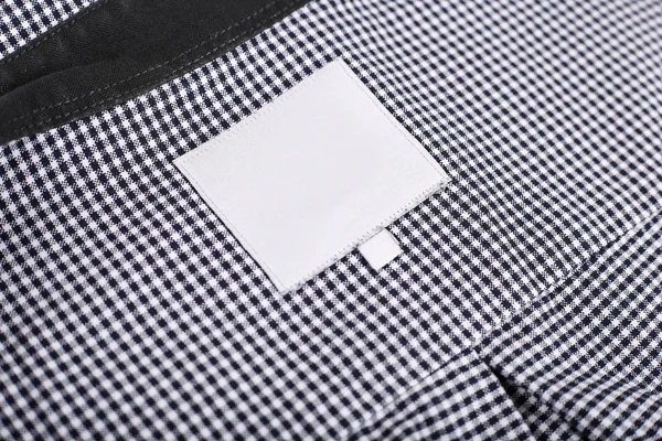 Vestiti in gabbia. Primo piano dell'etichetta dei vestiti bianchi su sfondo tessile. etichetta vestiti bianchi mockup  - — Foto Stock