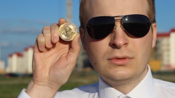 Ein Mann mit schwarzer Brille hält einen Bitcoin in der Hand und ist traurig, traurig, lockig — Stockvideo