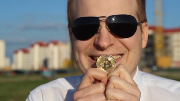 Um homem de óculos pretos está segurando uma bitcoin em suas mãos e rindo, feliz, close-up — Vídeo de Stock