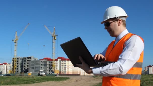 Inspector in black glasses makes data in a laptop on the background of the construction of a residential house — Stock Video