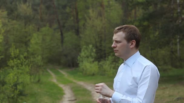 En man i en vit skjorta dricksvatten från en flaska i parken, törst — Stockvideo