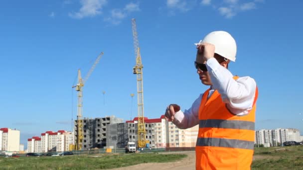 Inspektör boss i svarta glasögon på bakgrunden av byggandet av huset tar bort vita hjälmen från huvudet och torkar svetten — Stockvideo