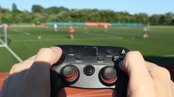 En man med en spelet joystick som spelar fotboll, i bakgrunden av stadion med riktiga människor som spelar fotboll — Stockvideo