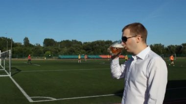 Bir adam siyah gözlüklü nerede futbol, futbol maçı ve içki bira oynamak stadyum içinde belgili tanımlık geçmiş bir bardak bira içiyor