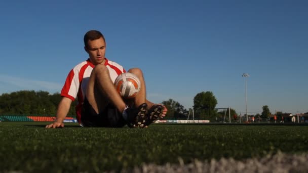 Spieler sitzt auf dem Rasen und hüpft mit den Füßen einen Fußball — Stockvideo