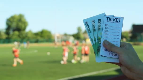Man håller en bookmakers biljett och pengar euro i bakgrunden av en stadion fotboll spel, närbild — Stockvideo