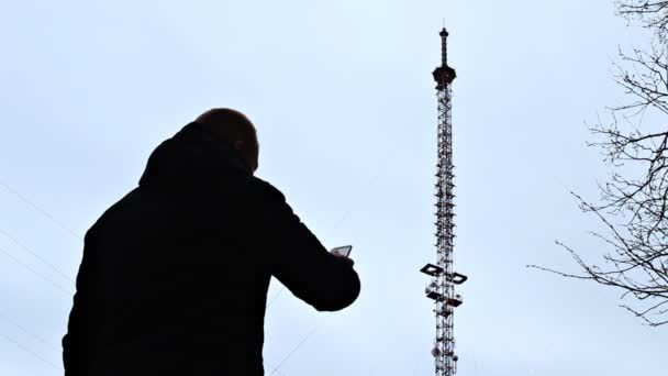 Un hombre con un teléfono está parado junto a la torre telefónica marcando un número de teléfono y tratando de conectar la red, una mala conexión, una señal débil — Vídeo de stock