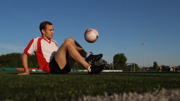 Игрок, сидящий на траве и прыгающий футбольный мяч ногами — стоковое видео