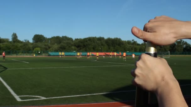 En man öppnar en flaska öl på bakgrunden av en fotbollsplan där de spelar fotboll, närbild — Stockvideo