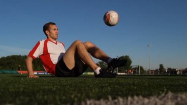 Çimenlerin üzerinde oturan ve bir futbol topu ayaklar tarafından sıçrayan oyuncu