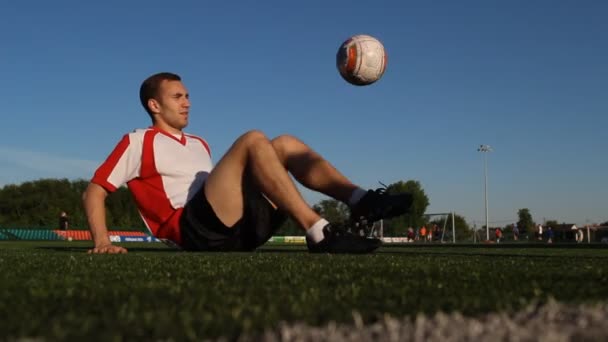 Игрок, сидящий на траве и прыгающий футбольный мяч ногами — стоковое видео