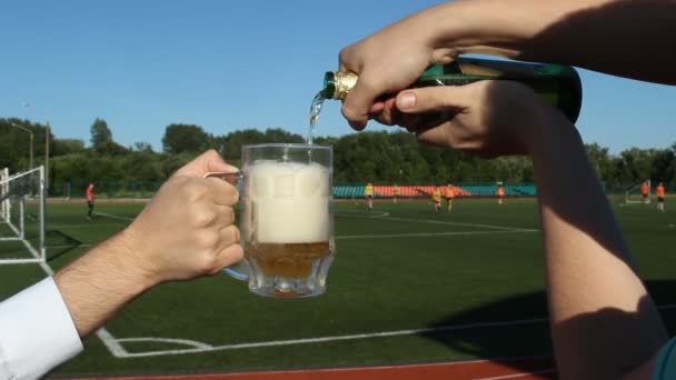 Un homme verse de la bière dans un verre sur le fond d'un stade de football où ils jouent au football, gros plan — Video