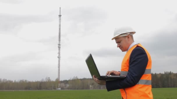 A man in a helmet inspector with a laptop records the data checks and looks toward the telephone tower — Stock Video