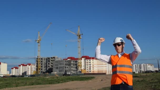 Der Ingenieur mit der schwarzen Brille und der Signalweste jubelt vor dem Hintergrund des Hausbaus — Stockvideo