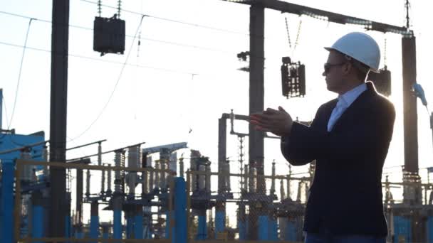 Un homme avec un casque blanc et des lunettes de soleil se frotte les mains sur le fond d'une centrale électrique, le coucher du soleil — Video