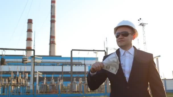 Un joven inspector de ingeniería satisfecho en gafas negras con dólares en dinero contra el fondo de una planta de energía — Vídeos de Stock