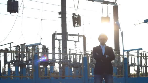 Ingeniero hombre en un casco blanco y gafas negras se levanta contra el telón de fondo de una central eléctrica, sus brazos cruzados, puesta de sol — Vídeos de Stock