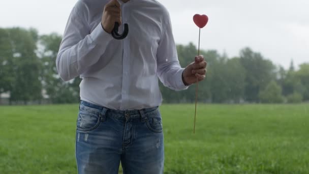 Un hombre está de pie bajo un paraguas, su lluvia y girando en sus manos un corazón rosado, la homosexualidad, el concepto de la LGBT, slow-mo — Vídeo de stock