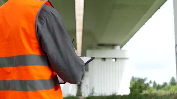 L'ispettore registra le violazioni riscontrate durante l'ispezione del ponte attraverso il fiume, primo piano, esaminatore e ponte — Video Stock