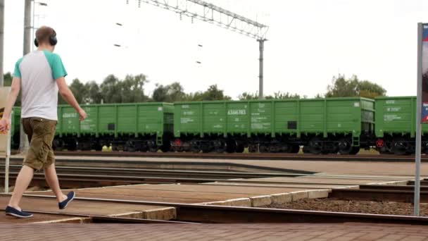 Een jonge man in hoofdtelefoons kruist het spoorlijnen en luistert naar de muziek, de naderende trein niet te horen — Stockvideo