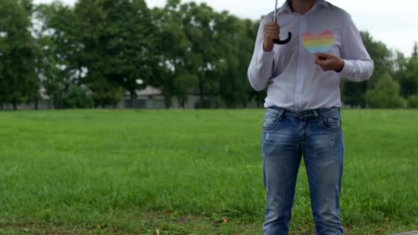 Un hombre con una camisa blanca está de pie bajo un paraguas está lloviendo y sosteniendo su corazón en forma de bandera LGBT, homosexualidad — Vídeo de stock