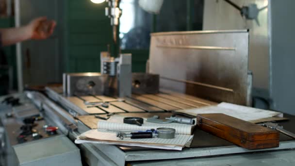 Un taller privado para trabajar con piezas metálicas, en el fondo una máquina de perforación perfora un agujero en la polea, el hombre toma las herramientas de medición de la mesa, la mano — Vídeos de Stock