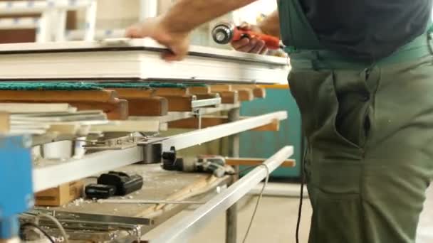 Produzione e produzione di finestre in pvc, un lavoratore maschio assembla una finestra profilo in pvc, trivella un foro nel profilo in pvc per il montaggio di parti incernierate, trapano finestra in pvc — Video Stock