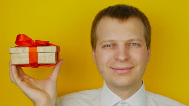 Hombre con regalo dentro de la caja sonríe y mira a la cámara, sobre fondo amarillo de la pared — Vídeos de Stock