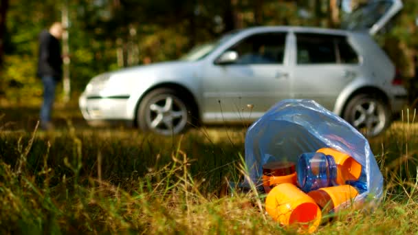Un paquete de basura con botellas de plástico se encuentra en la naturaleza, en el fondo hay un coche y la gente está caminando, la contaminación de la naturaleza con residuos de plástico, un coche en la naturaleza y la basura, basura — Vídeos de Stock