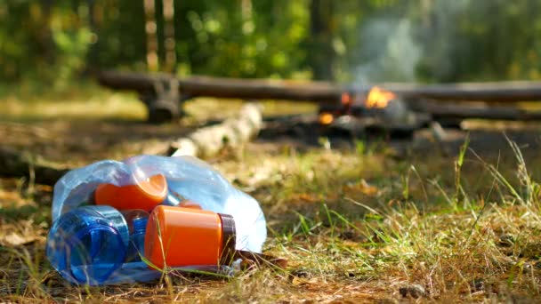 Человек бросает пластиковую бутылку в природе, огонь горит на заднем плане, открытый отдых, природа и мусор, кемпинг — стоковое видео