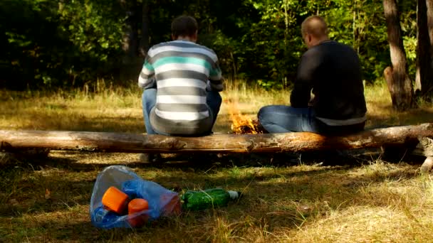 Ein Paket mit Plastikmüll liegt auf einer Lichtung, ein Feuer brennt, Menschen laufen im Hintergrund, Camping- und Naturverschmutzung, Müll — Stockvideo