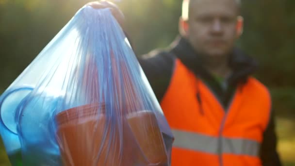 Un hombre en un chaleco naranja de señal sostiene un paquete con basura en el fondo de la naturaleza, bosque, primer plano, recolección de basura, un conserje, voluntario — Vídeos de Stock