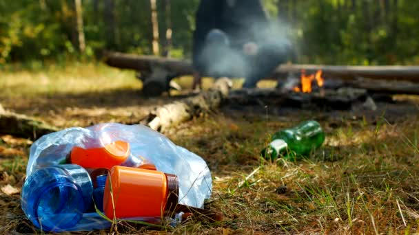 Пакет з пластиковим сміттям лежить на галявині, пожежа горить, люди ходять на задньому плані, кемпінг та забруднення природи, сміття — стокове відео