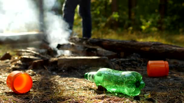 In the foreground in a forest glade there are plastic bottles of garbage, fires are smoking, people walk in the background, outdoor recreation, pollution by garbage — Stock Video