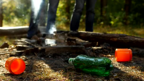 На передньому плані в лісовій галявині є пластикові пляшки сміття, пожежі курять, люди ходять на задньому плані, відпочивають на вулиці, забруднення сміттям — стокове відео