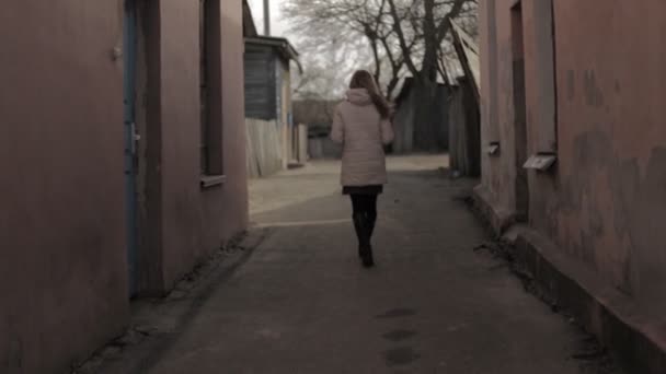 En tjej i en jacka med långt hår går längs en smal urban gränd, urban, långsamma-mo, smala gränd — Stockvideo