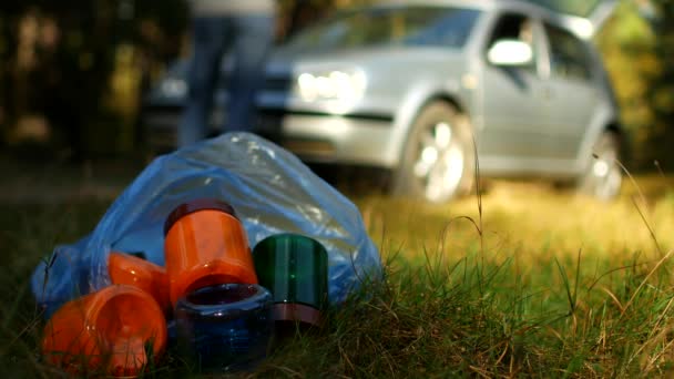 Пакет сміття з пластиковими пляшками лежить на природі, на задньому плані автомобіль і люди ходять, забруднення природи пластиковими відходами, автомобіль на природі і сміття — стокове відео