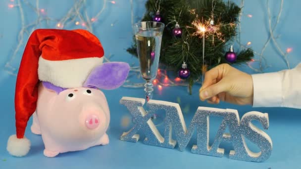 Мягкая игрушечная розовая свинья в красной шляпе Санта-Клауса на фоне елки на синем фоне, бокал шампанского, мужчина зажигает искру, копирует пространство, замедленная съемка — стоковое видео