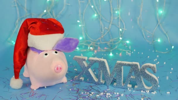 Plüschtier rosa Schwein mit rotem Weihnachtsmann-Hut auf dem Hintergrund des Weihnachtsbaums auf blauem Hintergrund, die Aufschrift Weihnachten, die Explosion der Weihnachtcracker und Weihnachtslametta und Bonbons — Stockvideo