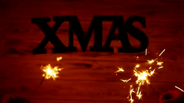 Inscription de Noël XMAS balancent librement dans l'air, sur un fond en bois blanc, étincelles brûlent en arrière-plan, Fêtes du Nouvel An, Noël, fond — Video