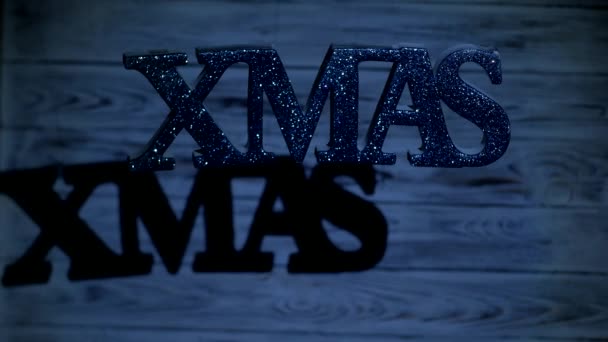 Inscripción de Navidad XMAS cuelga libremente en el aire, sobre un fondo de madera blanca, sobre la que brilla un haz de luz de la linterna, las vacaciones de Año Nuevo, el nuevo año 2019, hristmas — Vídeo de stock