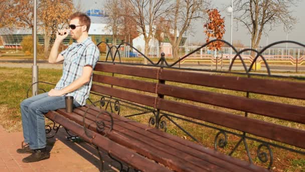 Fashion man s'assoit sur un banc, fume une cigarette électronique et boit du café — Video