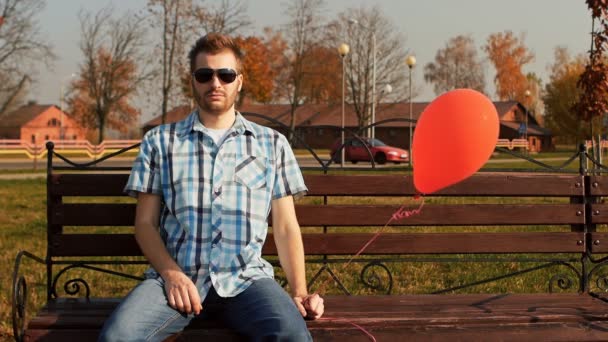Σοβαρή γενειοφόρος άντρας κάθεται σε ένα παγκάκι με ένα κόκκινο μπαλόνι στο χέρι του — Αρχείο Βίντεο