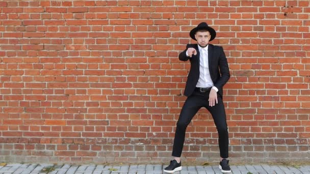 Un jeune homme caucasien vêtu d'un costume et d'un chapeau noirs danse et s'amuse contre un mur de briques, ralentit, copie l'espace — Video