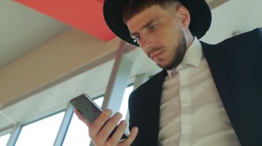 Bir çanta ve şapkası sakallı şık beyaz erkek işadamı duran ve iş merkezi, yakın çekim, Güneş, ağır çekim bir akıllı telefon bilgileri okuma