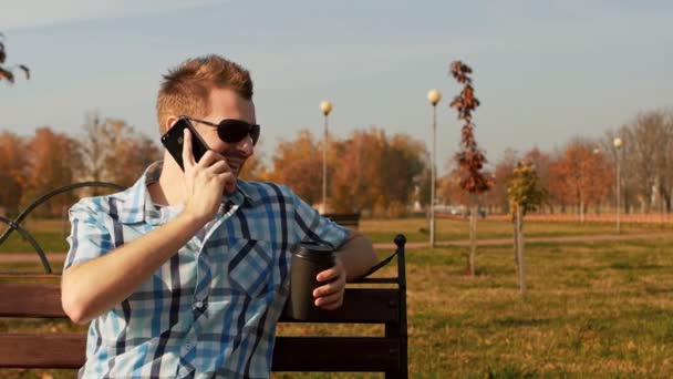 Brodaty facet z filiżanką kawy siedzi na ławce i rozmowy na telefon — Wideo stockowe