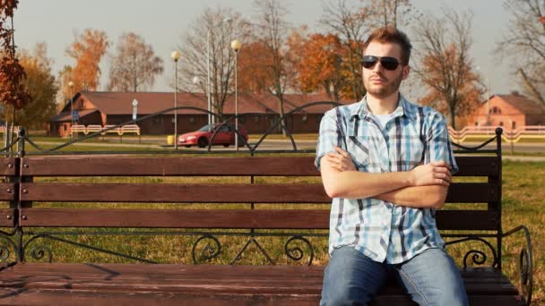 Eleganta skäggiga killen i solglasögon klädd i skjorta och jeans sitter på en bänk — Stockvideo