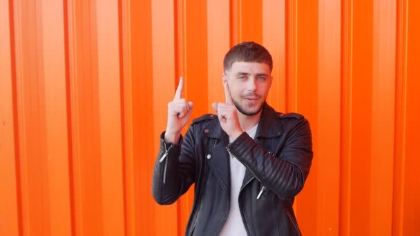 Young caucasian cheerful brunette man in black leather jacket is dancing on orange background, close-up, slow motion, outdoor — Stock Video