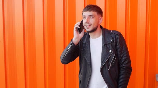 Молодой небритый кавказский мужчина в черной куртке разговаривает по телефону на оранжевом фоне, медленно, модно — стоковое видео
