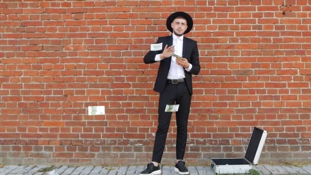 Un jeune homme en costume noir et chapeau compte des dollars d'argent sur fond de mur de briques, ralenti, style — Video